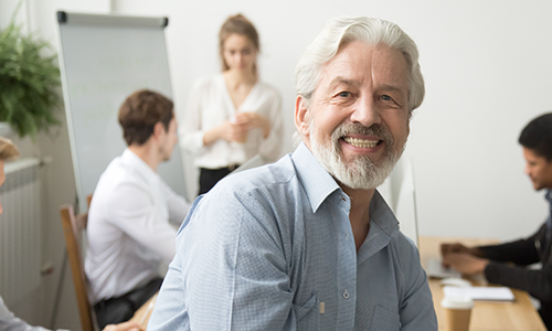 An older businessman poses for a portrait with a team brainstorming on laptops and an easel in the background.