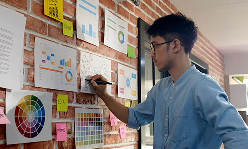 Young designer drawing a bubble map on a paper taped to a brick wall. Many other papers with charts and colors are also taped to the wall.