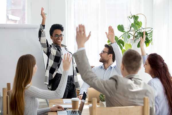 Diverse group of colleagues sit around a conference table, happily raising their hands as directed by the facilitators who is in the front of the room by a blank easel.