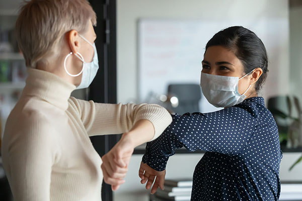Diverse colleagues wear face masks and touch elbows to greet each other in an office.