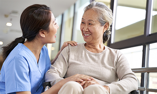A smiling female caregiver kneels down to create eye contact with a smiling older female patient in a wheelchair.