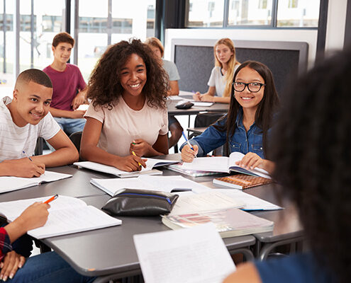 Diverse, happy high school students sit around a classroom table ready to take notes while listening to a speaker at the front of the classroom.