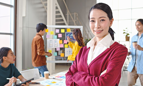 Portrait of a business woman smiling while a team of creatives meets in the background, using sticky notes and tablets for brainstorming.
