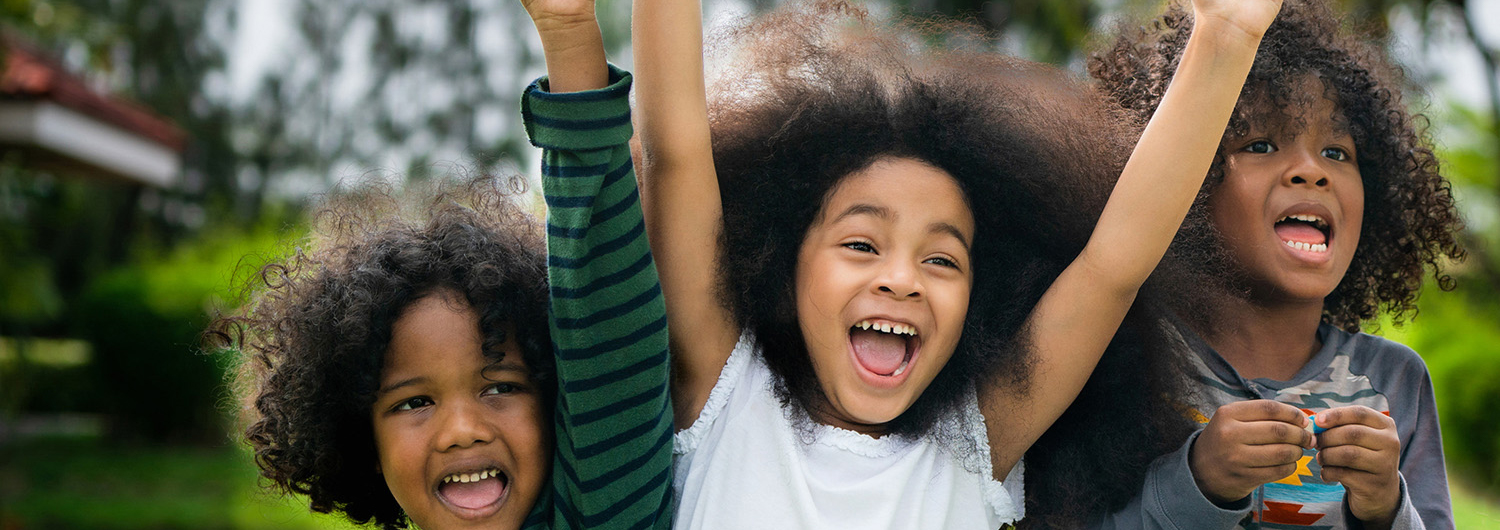 Close up of three diverse, happy children, two with their arms up in the air in excitement.