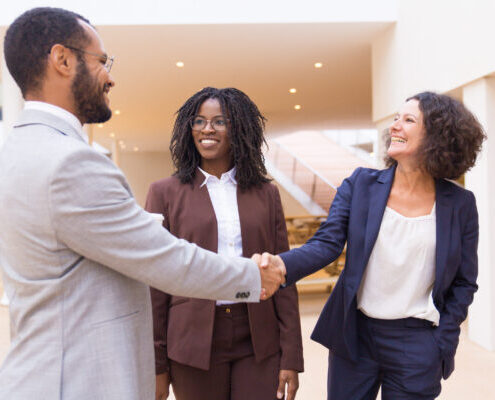 Diverse happy business partners wearing formal business attire greet each other by shaking hands in a lobby area.