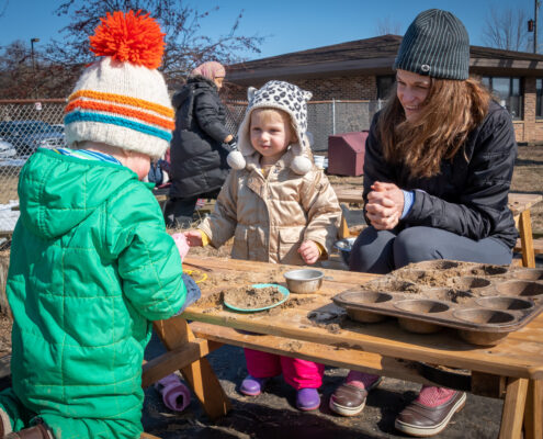 Toddlers playing with an adult in a mud kitchen.