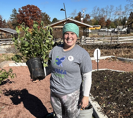 In a park setting, Lindsey Engelhardt holds up a gallon size bushy plant beside a cleared out garden on a chilly fall day.