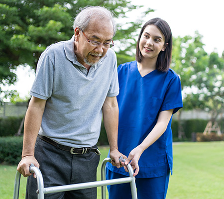 An older male walks outside, assisted by his walker and a young nurse.