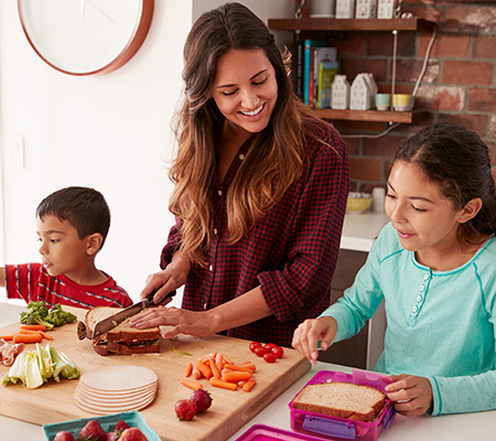A mother and two children prepare lunch containers with a sandwich and fresh fruits and veggies.