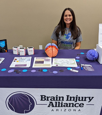 Jennifer Kirchen smiles sitting behind a table spread with information on brain injuries.