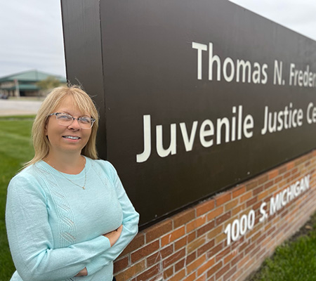 Suzanne West stands smiling outside by a building sign for the Thomas N. Frederick Juvenile Justice Center.